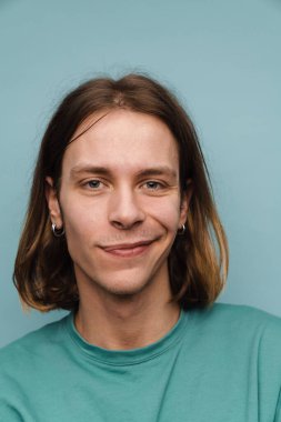 Tişört giyen genç beyaz adam gülümsüyor ve mavi duvarın üzerinden izole edilmiş kameraya bakıyor.