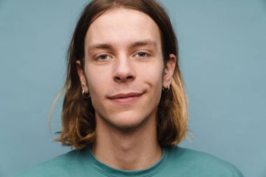 Tişörtlü gülümseyen genç sarışın bir adamın portresi mavi arka planda kameraya bakıyor.