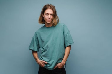 Tişört giyen genç beyaz adam gülümsüyor ve mavi duvarın üzerinden izole edilmiş kameraya bakıyor.