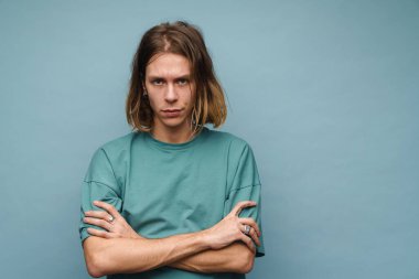 Genç beyaz adam kameraya bakarken kolları çaprazlanmış bir şekilde mavi duvarda poz veriyor.