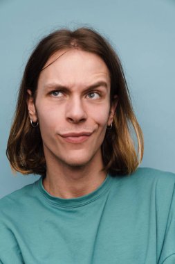 Tişört giyen genç beyaz adam mavi duvarın üzerinden yukarı bakarken kaşlarını çatıyor.