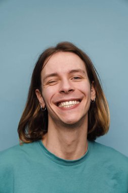 Genç beyaz adam tişört giyiyor gülümsüyor ve mavi duvarın üzerinden kameraya göz kırpıyor.