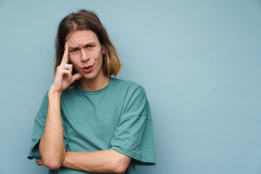 Mavi duvardan izole edilmiş kameraya bakarken kaşlarını çatan genç beyaz bir adam.