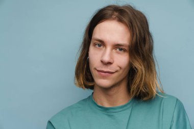 Tişört giyen genç beyaz adam gülümsüyor ve mavi duvarın üzerinden izole edilmiş kameraya bakıyor.