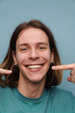 Tişört giyen genç beyaz adam gülümsüyor ve mavi duvarın üzerinde izole edilmiş dudaklarını işaret ediyor.