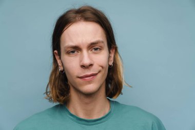 Tişört giyen genç beyaz adam gülümsüyor ve mavi duvarın üzerinden izole edilmiş kameraya bakıyor.