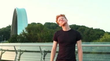 Yakışıklı kızıl saçlı bir adam köprüde dikilirken ısınma egzersizleri yapıyor.