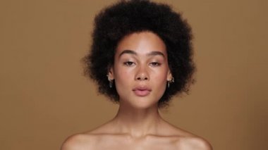 Muhteşem yarı çıplak Afro-Amerikalı bir kadın kameraya poz veriyor. Stüdyoda bej arka planda tek başına duruyor.