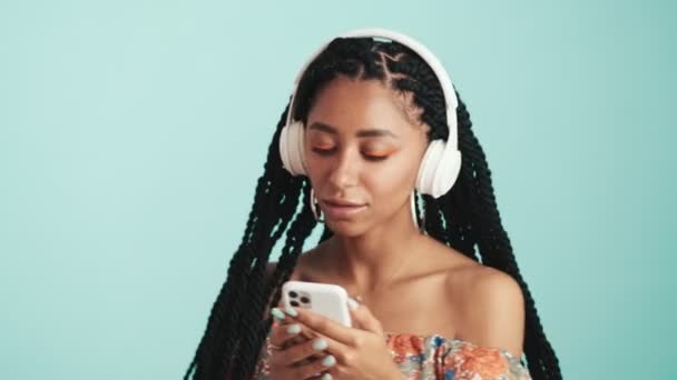 Симпатичная Африканка Слушает Музыку Наушниках Держа Телефон Изолированным Синем Фоне — стоковое видео