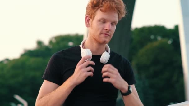 外を歩いている間に音楽をオンにするために彼のスマートウォッチを使用してヘッドフォンを持つ正の赤頭の男 — ストック動画