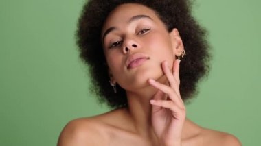 Yüzüne dokunurken poz veren yarı çıplak, Afro-Amerikalı çekici bir kadın stüdyoda açık yeşil arka planda tek başına duruyor.