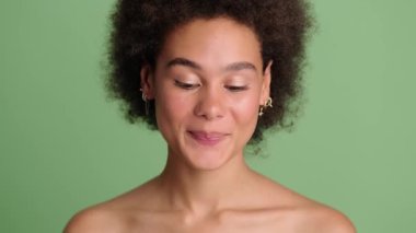 Mutlu, yarı çıplak, gülümseyen Afro-Amerikan bir kadın stüdyoda açık yeşil arka planda tek başına duruyor.