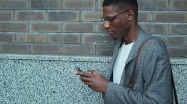 Memnun bir Afro-Amerikan işadamı binanın dışında dikilirken cep telefonuyla yan tarafa bakıyor.