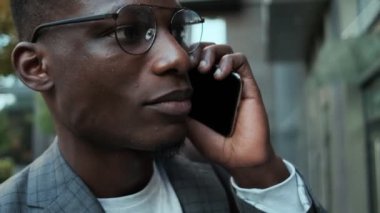 Yakışıklı bir Afro-Amerikan iş adamının binanın önünde dikilirken cep telefonuyla konuştuğu yakın plan bir görüntü.