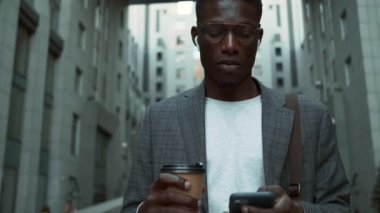 Sokakta yürürken kahve içerken telefonuyla kulaklık takan Afro-Amerikalı yakışıklı bir iş adamının yakın çekimi.