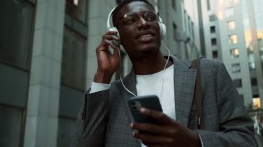 Memnun bir Afro-Amerikan iş adamının yakından görüntüsü. Kulaklıklı, cep telefonuyla konuşuyor. Dışarıda sokakta yürüyor.
