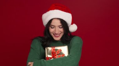 Yeşil kazak giyen ve Noel Baba şapkası takan pozitif bir kadın stüdyoda kırmızı arka planda izole bir şekilde duran hediye kutusuyla poz veriyor.