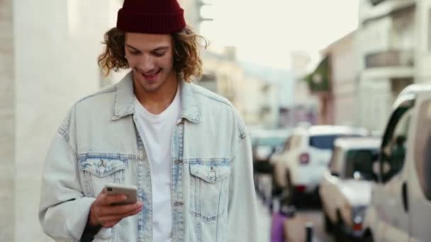 Egy Boldog Férfi Farmerdzsekiben Nézi Telefonját Miközben Sétál Gördeszkát Cipel — Stock videók