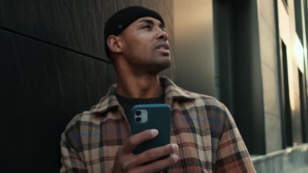 一位面带微笑的非洲裔美国人 身穿格子衬衫 拿着手机站在大楼外的一侧 — 图库视频影像