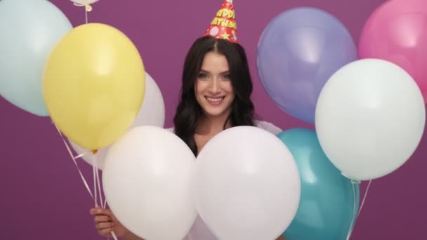 在演播室里 一个头戴一顶帽子 头戴气球的积极的生日女孩与紫色背景隔离地站在一起 — 图库视频影像