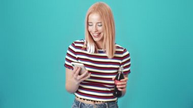 Kulaklıklı mutlu bir kadın stüdyodaki mavi duvarın üzerinde durmuş kola içerken cep telefonunu kullanıyor.