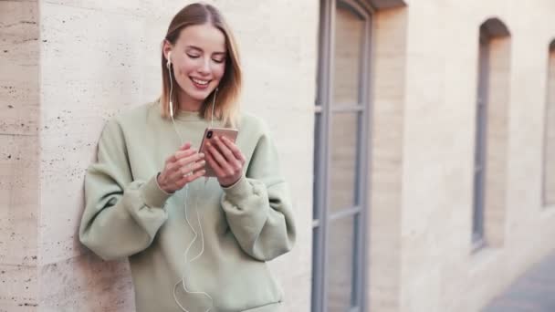 Happy Blonde Woman Listening Music While Using Her Earphones Standing — Stock Video