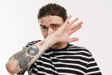 Genç beyaz adam yüzünü kapatıyor ve beyaz arka planda izole edilmiş kameraya bakıyor.