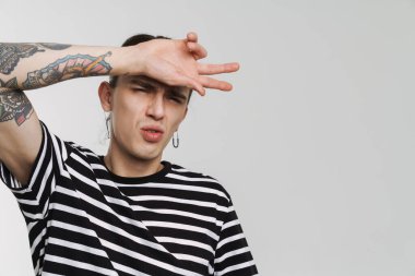 Küpeli genç beyaz adam beyaz arka planda izole edilmiş kameraya surat asıyor ve el kol hareketi yapıyor.