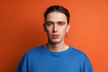 Küpeli genç beyaz adam poz veriyor ve turuncu arka planda izole edilmiş kameraya bakıyor.