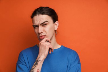 Küpeli genç beyaz adam somurtuyor ve turuncu arka planda izole edilmiş kameraya bakıyor.