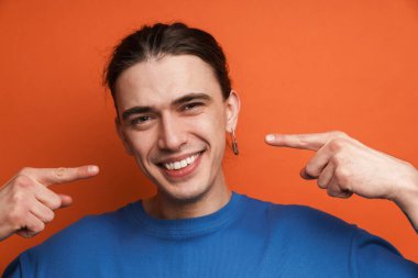 Küpeli genç beyaz adam gülüşünü işaret ediyor turuncu arka planda izole edilmiş.