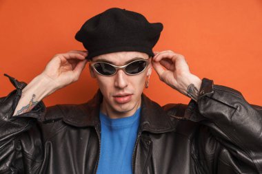 Deri ceketli genç beyaz adam turuncu arka planda kameraya bakıyor ve poz veriyor.