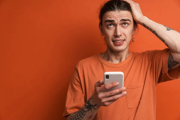 Young white man holding his head and using cellphone isolated over orange background