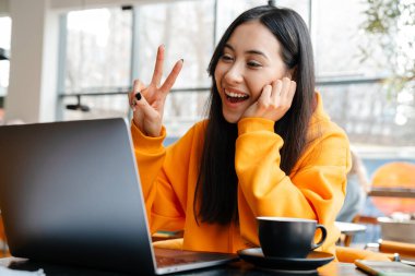 Gülümseyen Asyalı kadın barış işareti yapıyor ve kafede laptop kullanıyor.