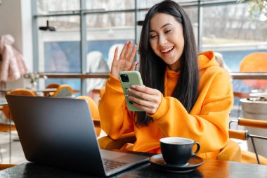 Kafede dizüstü bilgisayarla çalışırken cep telefonu kullanan gülümseyen Asyalı kadın.