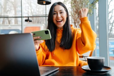 Heyecanlı Asyalı kadın internette cep telefonuyla oyun oynuyor ve kafede birinci sınıf jest yapıyor.