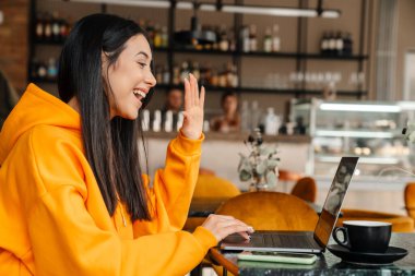 Gülümseyen Asyalı kadın kafede otururken diz üstü bilgisayar kullanıyor.