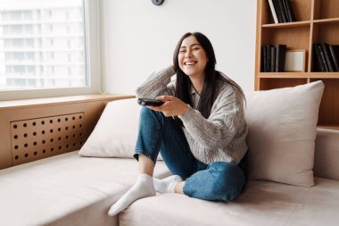 Evde kanepede oturmuş uzaktan kumandayla televizyon izleyen çekici genç bir kadın.