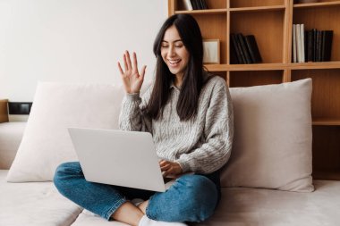 Kanepede oturmuş el sallayan ve evdeki dizüstü bilgisayardan vodeo çağrısı yapan çekici genç bir kadın.