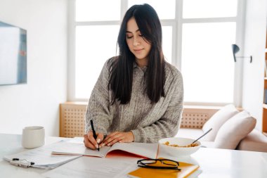 Çekici ve zeki bir kadın mutfakta sağlıklı bir kahvaltı yaparken ders çalışıyor.