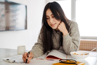Çekici ve zeki bir kadın mutfakta sağlıklı bir kahvaltı yaparken ders çalışıyor.