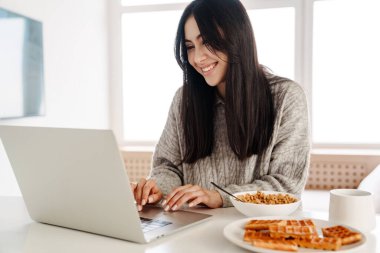 Genç ve çekici bir kadın kahvaltısını yaparken dizüstü bilgisayar kullanıyor.