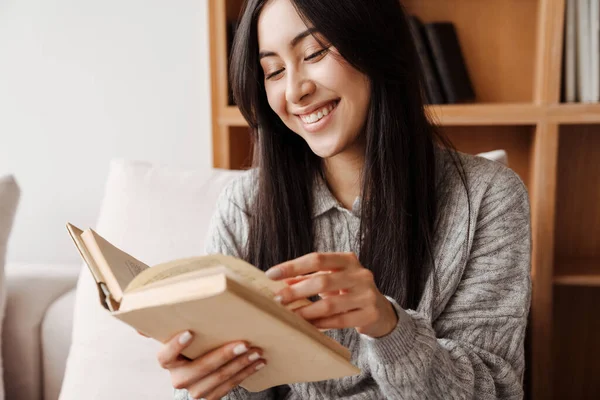 若いです幸せな女性読書本ながら座っています上のソファでザリビングルーム — ストック写真