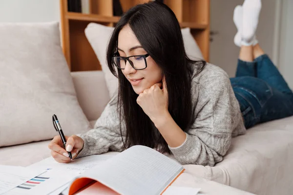 Feliz Asiático Estudiante Chica Haciendo Tarea Mientras Mintiendo Sofá Casa — Foto de Stock