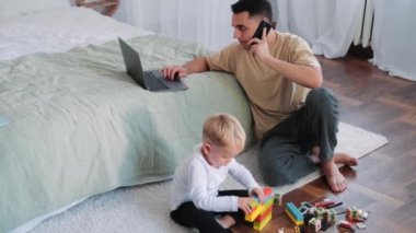 Küçük bir çocuk oyuncaklarıyla yerde oturuyor ve babası yanında oturuyor ve cep telefonuyla konuşuyor.