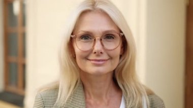 Sarışın, olgun bir iş kadınının, kameraya poz verip caddenin karşısında durduğu yakın plan görüntüsü.