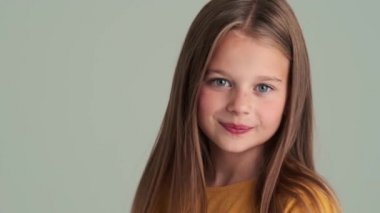 Stüdyonun gri duvarından izole edilmiş kameraya bakan utangaç küçük bir kızın yakın çekim görüntüsü.