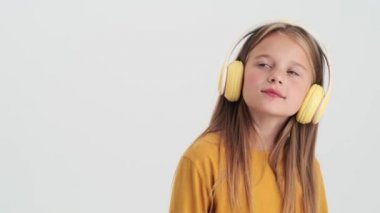 Sarı kulaklıklı mutlu küçük bir kız stüdyodaki gri bir duvarın üzerinde duran müziği dinliyor.