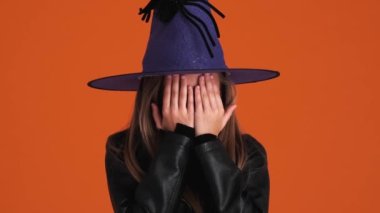 Cadı kostümü giymiş neşeli küçük bir kız elleriyle yüzünü örtüyor ve stüdyodaki turuncu bir duvarın üzerinde tek başına durduğunu gösteriyor.