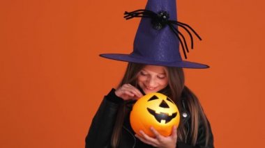 Cadı kostümlü mutlu küçük bir kız stüdyodaki turuncu bir duvarın üzerinde yapayalnız duran balkabağına bakıyor.